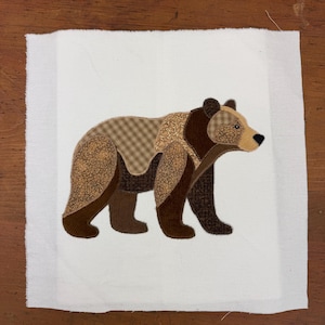 Puede incluir: Un parche de tela blanca cuadrada con una aplicación de oso hecha de varios textiles estampados en tonos marrón y beige. El oso se muestra de perfil, exhibiendo una mezcla de telas a cuadros, texturizadas y de color marrón sólido con costuras.