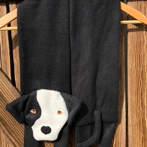Puede incluir: Bufanda de forro polar negro con una cara de perro blanco y negro y huellas de patas rosas en los extremos.