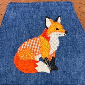 May include: A blue denim placemat featuring a patchwork fox design. The fox is composed of orange, white, and black fabric pieces, with a gingham pattern on the body. The fox is sitting, with its tail curled.