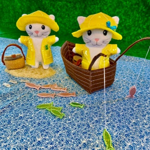 Pode incluir: Dois brinquedos de pelúcia de gato branco vestindo capas de chuva e chapéus amarelos. Um gato está segurando uma vara de pesca com um pequeno peixe na linha. Os gatos estão sentados em uma superfície com padrão azul e branco com vários peixes pequenos espalhados ao redor.