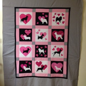 Könnte beinhalten: Eine rosafarbene und schwarze Steppdecke mit Patchwork-Muster, die verschiedene Hunderassen in Silhouette zeigt. Jedes Quadrat hat ein rosa Herz mit kleineren Herzen drumherum.