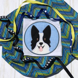 Könnte beinhalten: Ein weißer Stoffquadrat mit einem schwarzen und weißen gestickten Hundeface in einem blauen Kreis. Der Kreis ist mit Strasssteinen umrandet. Der Stoff liegt auf einem blau-grünen gemusterten Stoff.