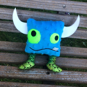May include: A blue plush monster toy with white horns and green scaly legs. The monster has large green eyes and a small mouth.