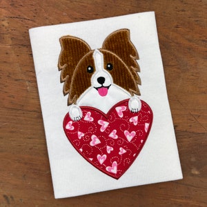 May include: A white t-shirt with an embroidered image of a brown and white papillon dog holding a red heart with pink hearts on it.