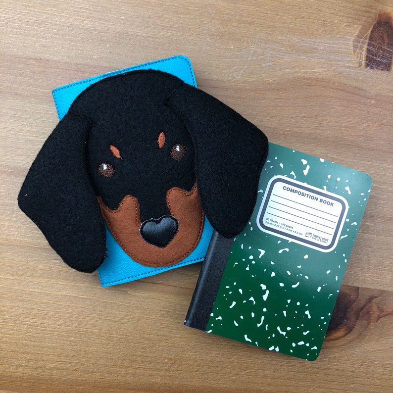 May include: A blue leather notebook cover with a black and brown dachshund dog face applique. A green and white composition notebook with the text "COMPOSITION BOOK" is next to the cover.