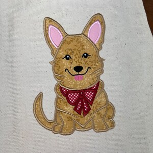 May include: Embroidered design of a brown dog with pink ears wearing a red and white checkered bow tie. The dog is sitting with its tongue sticking out.