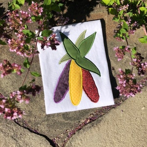 May include: A white t-shirt with an embroidered design of three corn cobs. The cobs are purple, yellow, and red, and the husks are green.
