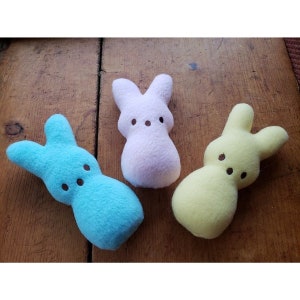 May include: Three plush bunny-shaped toys in pastel colors. The bunnies are light blue, light pink, and yellow. They have black eyes and a simple, smiling expression.