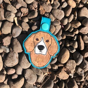 Puede incluir: Un llavero de corcho azul y marrón con la cabeza de un beagle bordada.