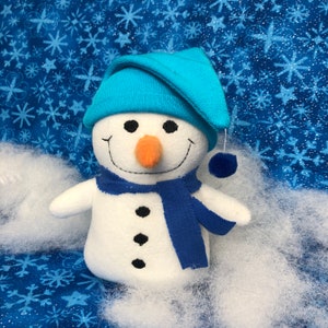 May include: A white plush snowman wearing a blue hat and scarf. The snowman has a carrot nose, black eyes, and a stitched-on smile. The snowman is sitting on a white fluffy surface against a blue background with white snowflakes.