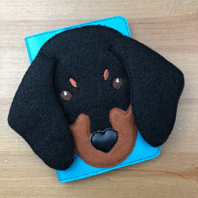 May include: A blue leather wallet with a black felt dachshund dog face applique. The dog has brown accents and a black heart-shaped nose. The wallet is on a wooden surface.