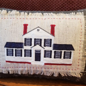 Op de afbeelding: Een decoratief kussen met een geborduurd wit huis op een beige linnen stof. Het huis heeft rode schoorstenen, zwarte ramen en een zwarte deur. Het kussen is omgeven door een donkerblauwe stof met een wit sterrenpatroon.