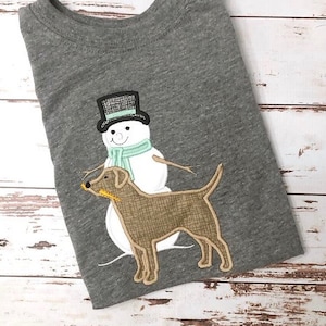 May include: A gray t-shirt with an embroidered design of a snowman wearing a black top hat and a light blue scarf, with a brown dog standing in front of the snowman.