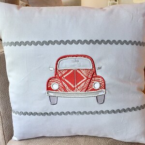 May include: A white pillow with a red embroidered Volkswagen Beetle design. The pillow has a gray scalloped trim around the edges.