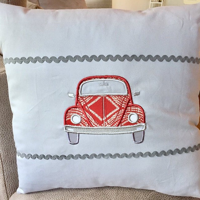 May include: A white pillow with a red embroidered Volkswagen Beetle design. The pillow has a gray scalloped trim around the edges.
