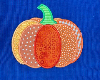 Puede incluir: Un aplique de calabaza colorido con secciones estampadas en naranja, amarillo y marrón, y un tallo verde. La calabaza está sobre un fondo de pana azul vibrante, ideal para la decoración de otoño.