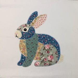May include: A patchwork bunny design featuring floral patterns in blue, pink, and beige. The bunny is depicted in a seated position, with its ears erect. The design is set against a white backdrop, highlighting the detailed stitching.