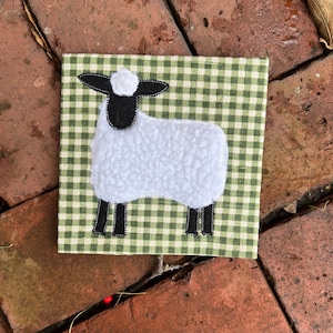 May include: A white sheep with black legs and a black face is appliqued on a green and white gingham fabric.