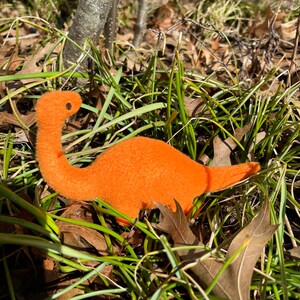 May include: A bright orange plush dinosaur toy with black eyes, lying in a bed of green grass and brown leaves.
