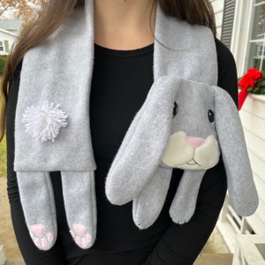 May include: A gray fleece scarf with a white pom-pom and a gray bunny face attached. The bunny has a white nose and pink cheeks.
