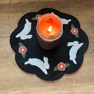 May include: A black felt candle mat with a white stitched design of bunnies and flowers. A glass candle with a white rim and a burning orange candle is in the center.