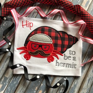 May include: A white t-shirt with an embroidered red and black plaid hermit crab wearing a white hat and glasses. The text "Hip to be a hermit" is embroidered below the crab.