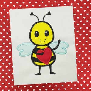Puede incluir: Una abeja de dibujos animados con rayas negras y amarillas que sostiene un corazón rojo. La abeja tiene una cara sonriente y alas azules.