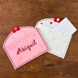 May include: Two felt envelopes with red heart buttons. The pink envelope has the name Abigail embroidered on it. The white envelope has a white card inside.