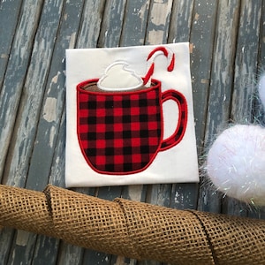 May include: Red and black plaid mug with whipped cream and a candy cane on top. The mug is embroidered on a white t-shirt.