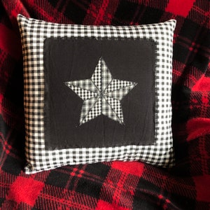 May include: A black and white checkered pillow with a black fabric center featuring a large white star. The pillow is sitting on a red and black plaid fabric.