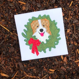 May include: Embroidered design of a dog's face peeking out of a green wreath with a red bow. The design is on a white fabric.