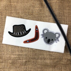 May include: A white fabric with three embroidered images: a brown cowboy hat, a brown boomerang, and a gray koala bear.