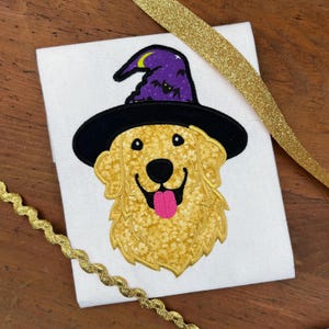 May include: A white t-shirt with an embroidered image of a golden retriever wearing a black witch hat with a purple pointed top and bats. The hat has a crescent moon and stars on the top.