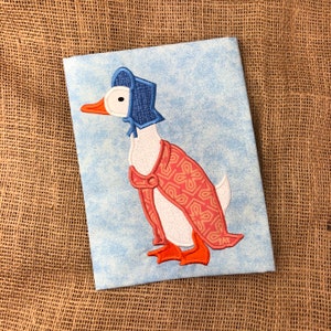 May include: A white duck with a blue bonnet and a red and orange patterned coat. The duck is embroidered on a light blue fabric.