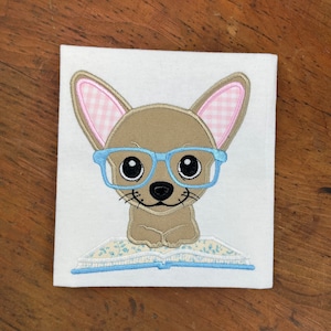 Puede incluir: Un chihuahua marrón con gafas azules y una diadema a cuadros rosa y blanca está sentado en un libro azul con flores blancas. El chihuahua está sonriendo y tiene una expresión linda.