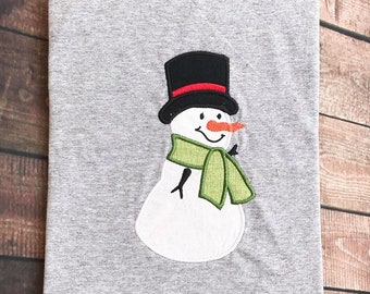 May include: A gray t-shirt with an embroidered snowman design. The snowman is wearing a black top hat with a red band, a green scarf, and has a carrot nose.