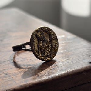 Op de afbeelding: Een bronskleurige ring met een cirkelvormig blad voorzien van een verhoogd, getextureerd reliëf van een figuur bij een boom. De metalen band splitst zich in twee dunne delen waar deze aansluit op het ronde middendeel, met een donkere patina.