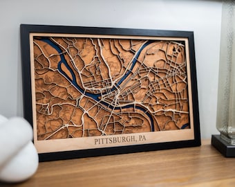 May include: A framed wooden map of Pittsburgh, Pennsylvania, with a blue river and streets carved into the wood. The map is in a black frame and has the text "PITTSBURGH, PA" at the bottom.