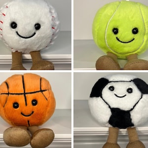 May include: Four plush toys shaped like sports balls: a baseball, a tennis ball, a basketball, and a soccer ball. Each toy has a smiling face and short legs.