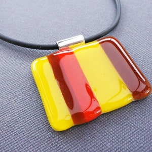 Op de afbeelding: Een ketting met een rechthoekige hanger van glas met gele, rode en bruine strepen. De hanger hangt aan een zwart koord. De hanger is ongeveer 4 cm breed en 2,5 cm hoog.