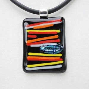 May include: A rectangular black glass pendant with colorful horizontal stripes in red, orange, yellow, and white. A small, iridescent blue and green oval shape is near the center. The pendant hangs from a black cord with a silver-tone metal clasp.