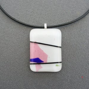Peut inclure: Un collier pendentif rectangulaire avec un fond blanc, des formes abstraites roses et bleues, et des lignes noires. Le pendentif est attaché à un cordon noir. Le pendentif mesure environ 3,8 cm de long et 2,5 cm de large.