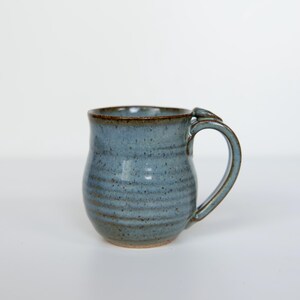 May include: A handmade blue ceramic mug with a brown rim and handle. The mug has a textured surface with horizontal lines and a rounded shape. The mug is designed for hot beverages.