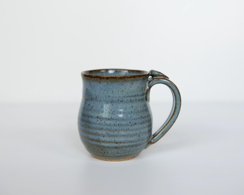 May include: A handmade blue ceramic mug with a brown rim and handle. The mug has a textured surface with horizontal lines and a rounded shape. The mug is designed for hot beverages.
