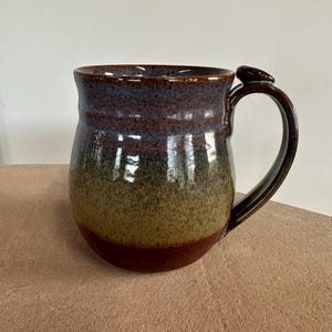 May include: A handmade ceramic mug with a rounded body and a dark brown handle. The mug features a speckled glaze with shades of brown, blue, and green. The mug is designed for hot beverages.
