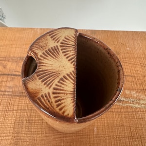 May include: A handmade ceramic utensil holder with a unique design. The holder is a mix of brown and tan colors, with a half-circle lid featuring a fan-like pattern. The interior is dark brown, and the exterior has a glossy finish.