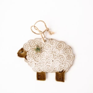 May include: A ceramic sheep ornament with a white body featuring a spiral design. The sheep has brown legs, a brown head, and a green star accent. A natural twine loop is attached for hanging. This rustic ornament is perfect for farmhouse decor.