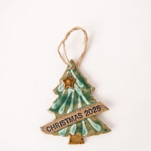 May include: A ceramic Christmas tree ornament with a star and the text "CHRISTMAS 2025". The ornament is green and brown with a jute string for hanging. A festive holiday decoration.