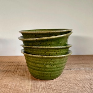 May include: A stack of four green ceramic bowls. The bowls have a textured, ridged exterior and a slightly flared rim. The bowls are a deep, earthy green color, with subtle variations in tone. They are stacked on a wooden surface.