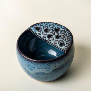 May include: A handcrafted, blue ceramic flower frog with a unique design. The bowl has a rounded shape with a textured, patterned top section. The color is a deep blue with brown accents.